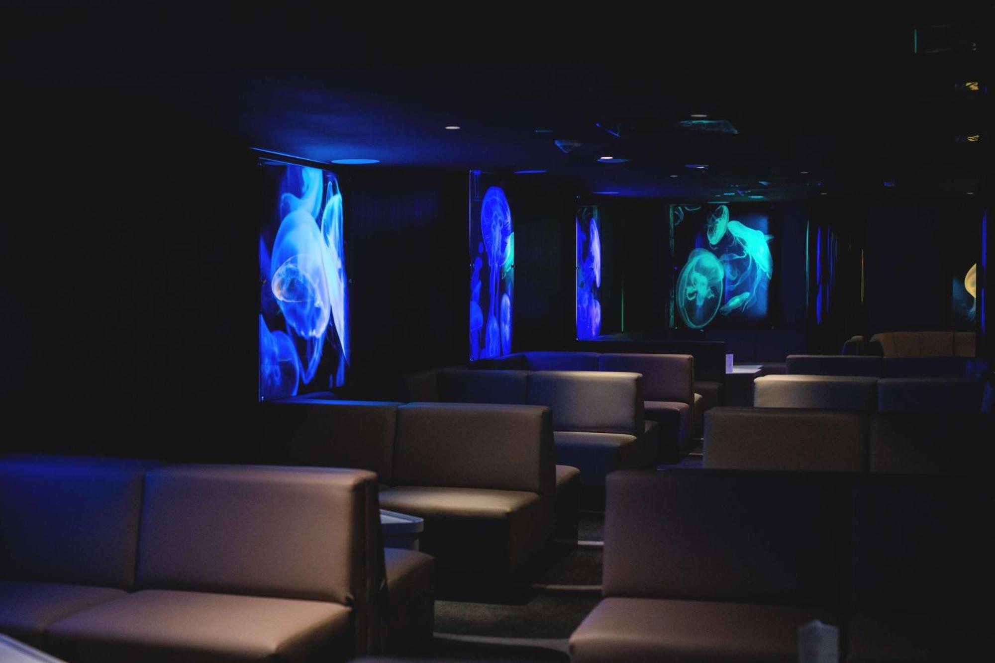 Radisson Blu Resort Swinoujscie Luaran gambar The photo shows a dimly lit interior space, likely a lounge or a seating area. The walls are adorned with glowing blue and turquoise images, possibly of jellyfish or abstract aquatic forms, creating an ambient atmosphere. There are several upholstere