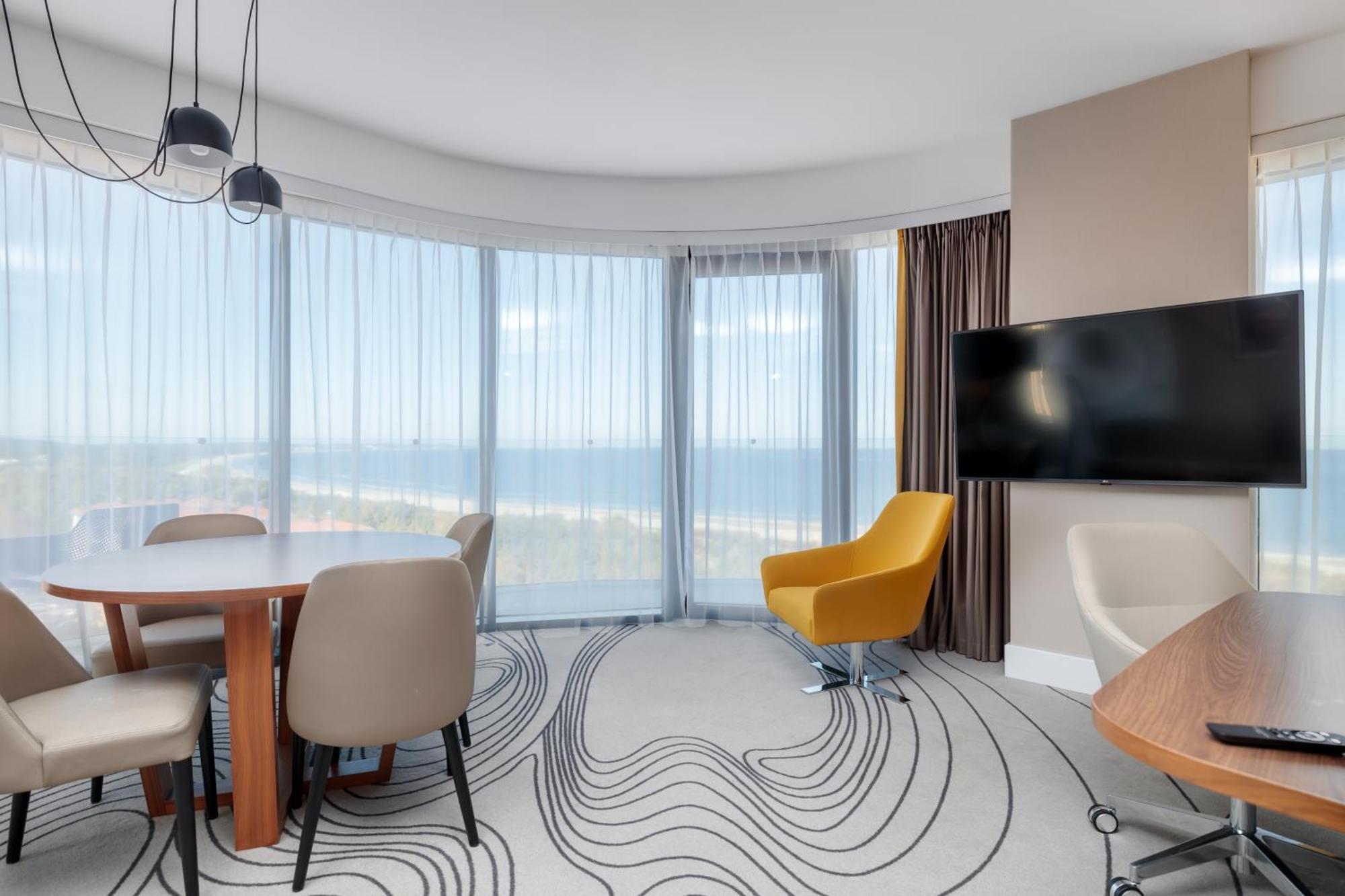 Radisson Blu Resort Swinoujscie Luaran gambar The image shows a modern hotel room or suite with a bright, airy atmosphere. There are large windows that allow natural light to fill the space, providing a view of the ocean. The decor includes a round dining table with several chairs, and a stylish