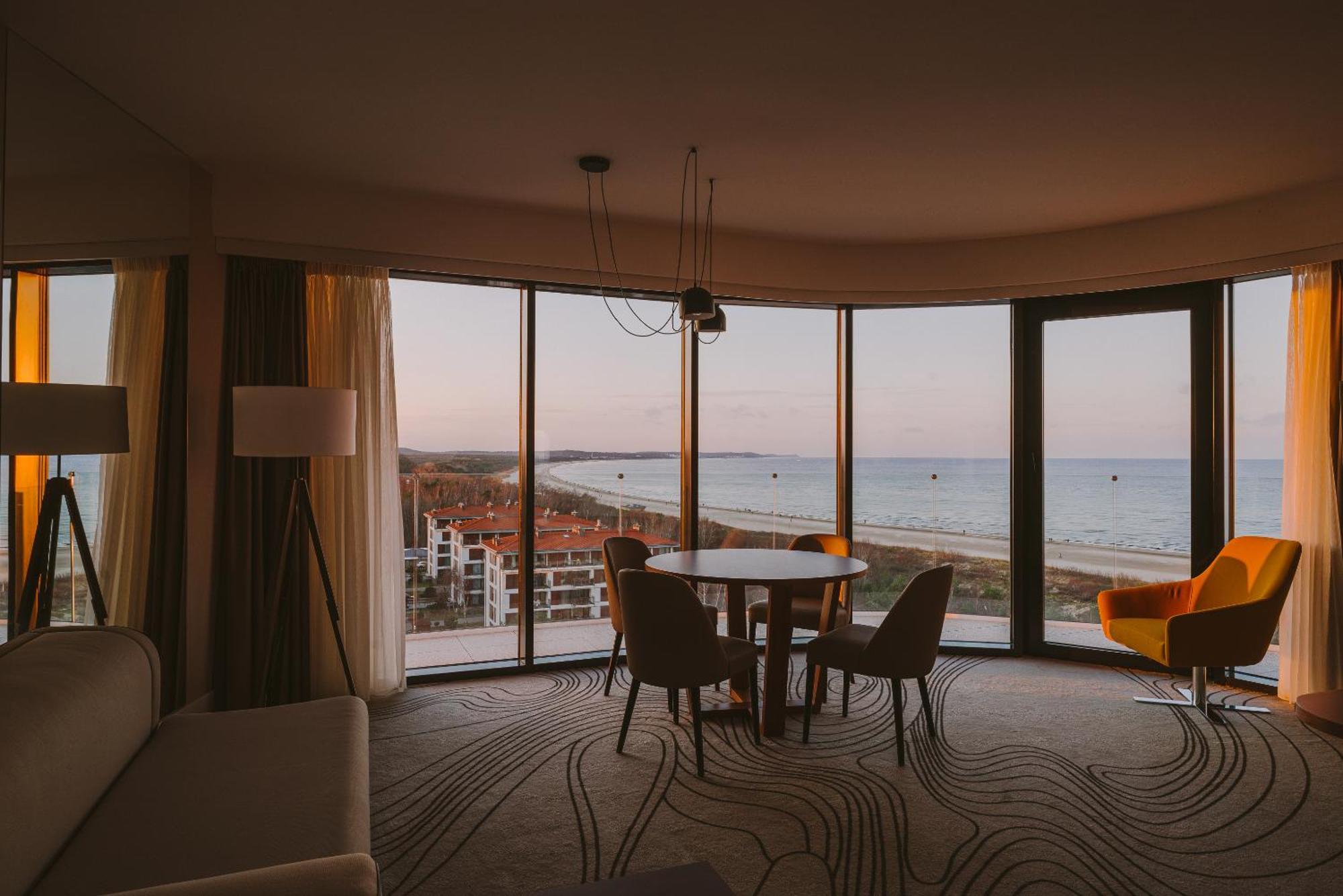 Radisson Blu Resort Swinoujscie Luaran gambar The photo shows a modern and stylish interior of a room featuring large windows that provide a view of the beach and ocean. The room is furnished with a round table and chairs, along with a cozy sofa and a bright yellow armchair. The decor has a cont