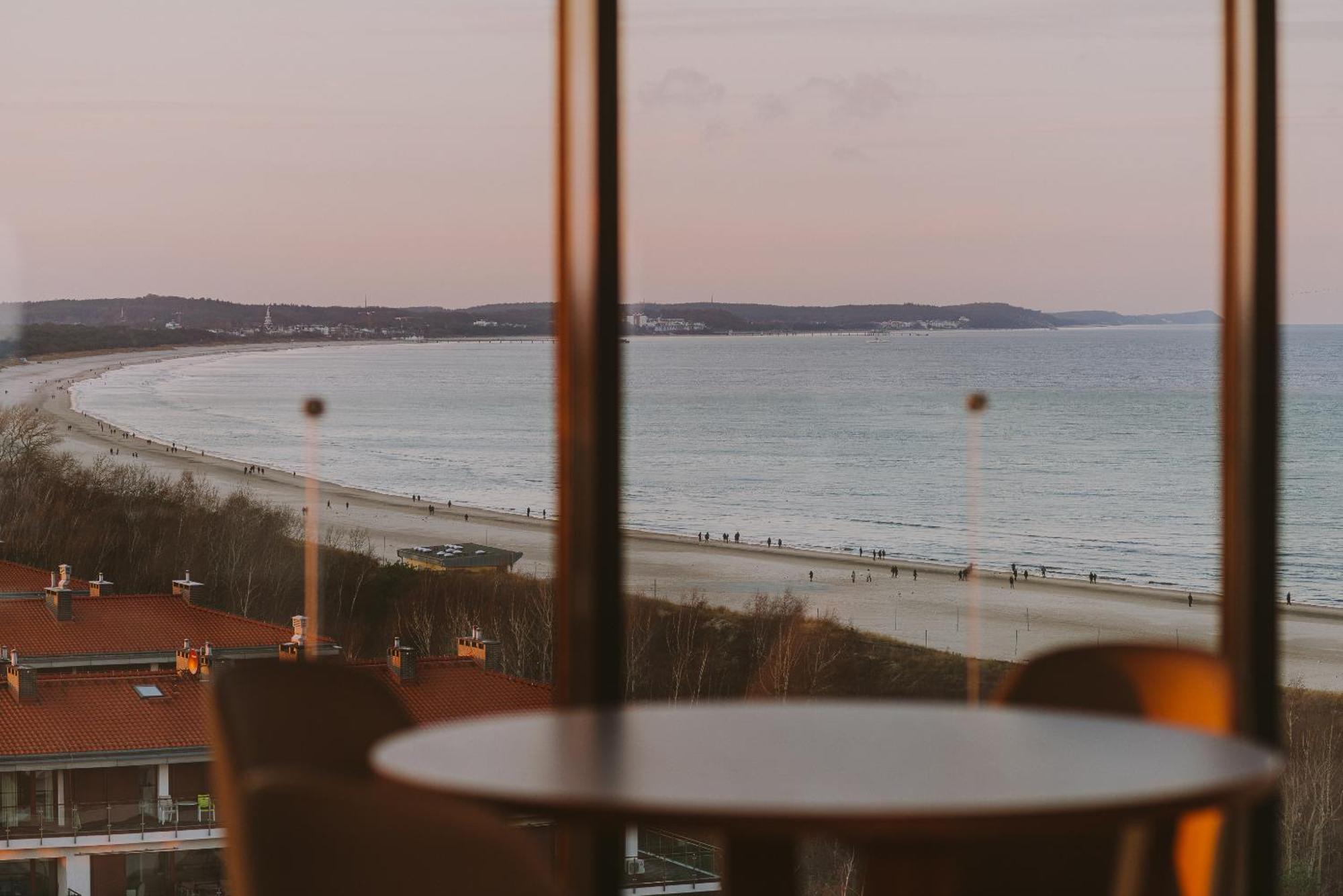Radisson Blu Resort Swinoujscie Luaran gambar The photo shows a picturesque view of a beach scene from indoors. In the foreground, there is a round table with some chairs around it, suggesting a cozy interior space. Through a large window, you can see the beach with gentle waves hitting the shor