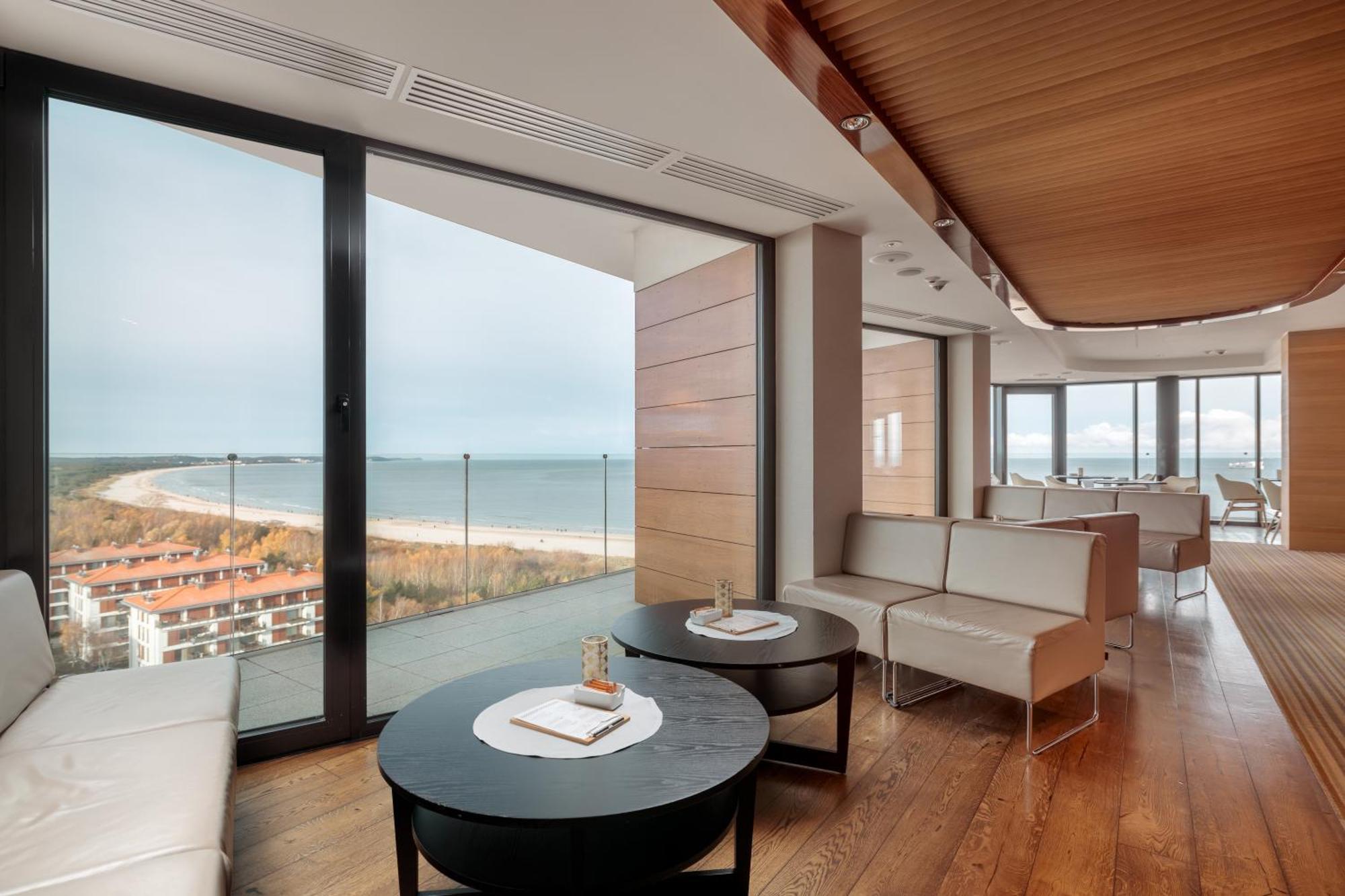 Radisson Blu Resort Swinoujscie Luaran gambar The photo shows a modern, elegantly designed interior space featuring large windows that offer a panoramic view of the ocean and coastline. The flooring is wooden, providing a warm atmosphere. There are two round black tables in the foreground with m