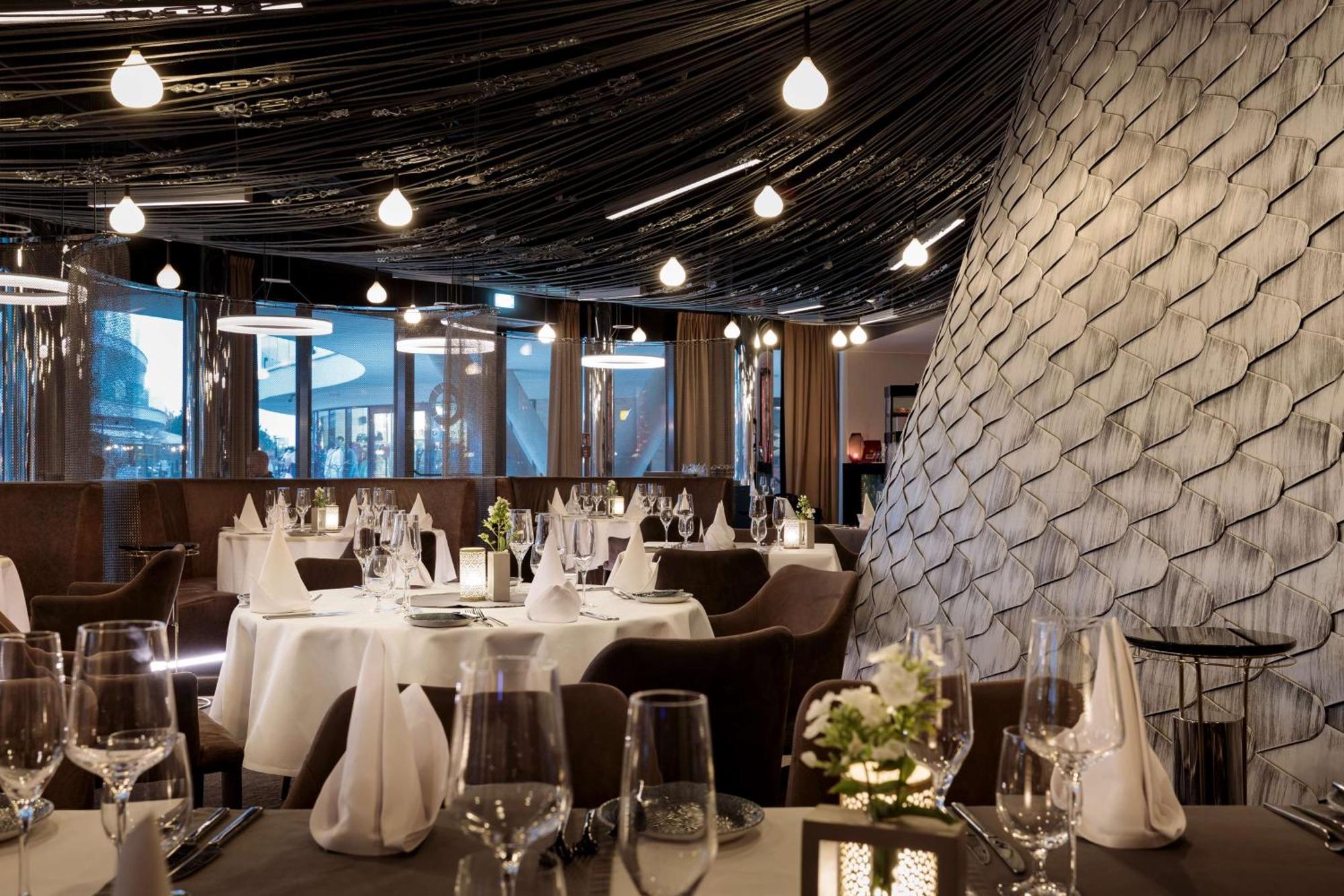Radisson Blu Resort Swinoujscie Luaran gambar The photo shows a stylish restaurant interior. The dining area features elegantly set tables with white tablecloths, glassware, and folded napkins. There are decorative elements like small floral arrangements on the tables. The ceiling is adorned wit