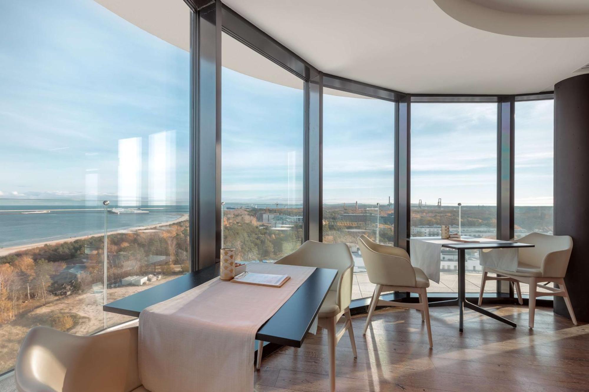 Radisson Blu Resort Swinoujscie Luaran gambar The photo shows a modern dining area with large windows that offer a panoramic view of the landscape outside, including a body of water and possibly a shoreline. The design features sleek furniture, including a table with chairs arranged around it, a