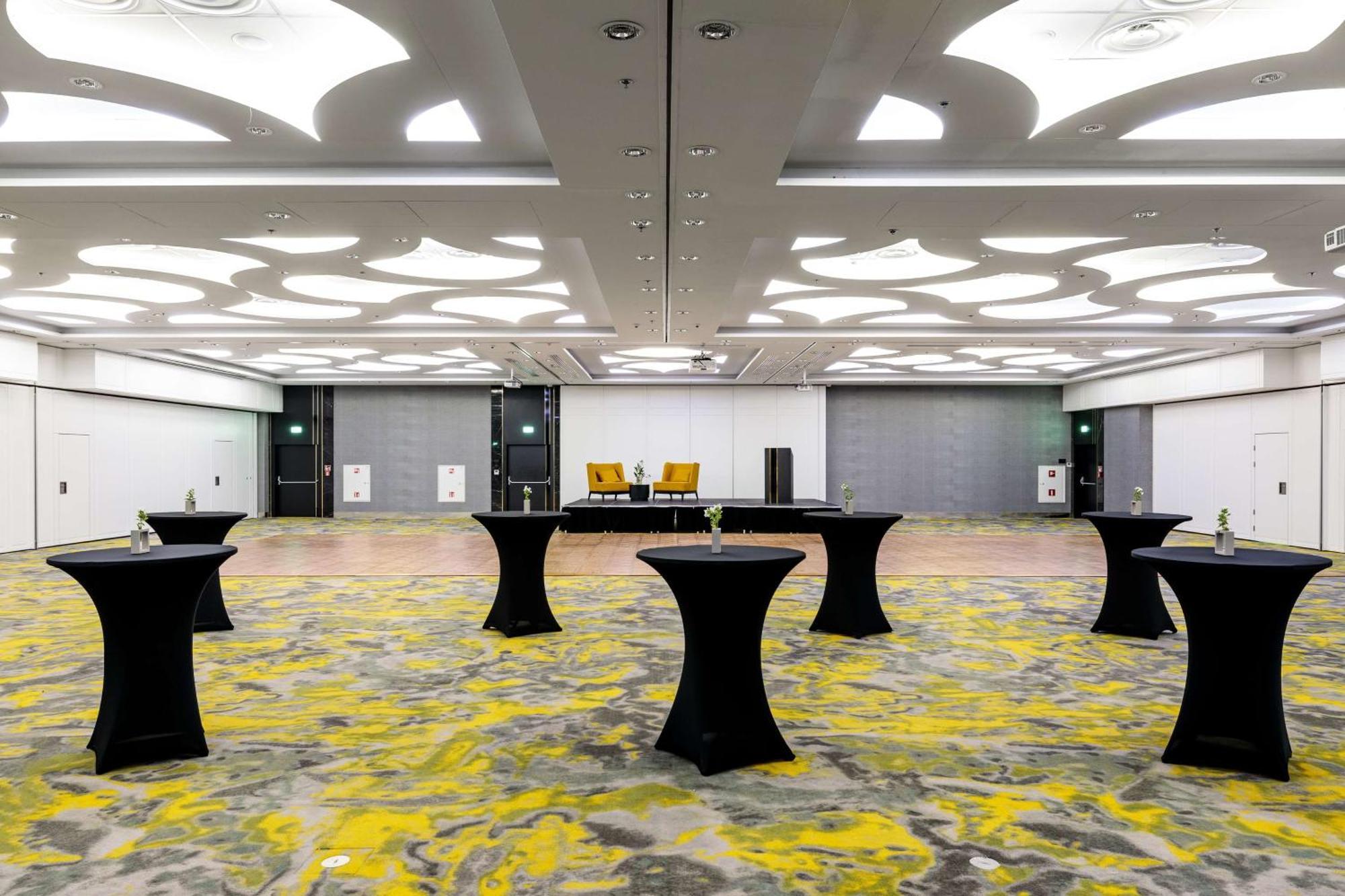 Radisson Blu Resort Swinoujscie Luaran gambar The photo shows a spacious interior room set up for an event. The flooring features a patterned design in gray with yellow accents. There are several black tall tables scattered throughout the area, and in the background, there are two large yellow a