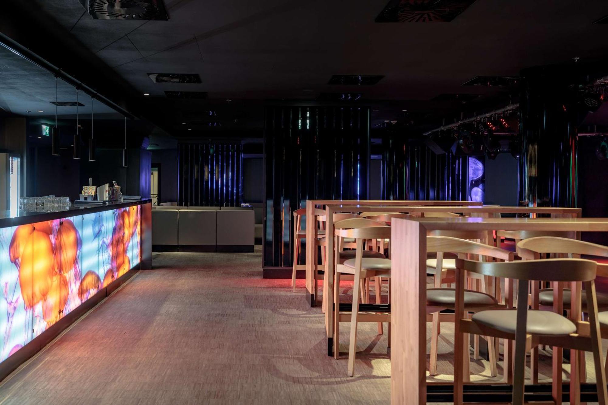 Radisson Blu Resort Swinoujscie Luaran gambar The photo shows the interior of a modern bar or nightclub. It features a sleek and stylish design with a long bar counter illuminated by colorful lights. The floor has a neutral carpet, and there are wooden high-top tables with accompanying chairs ar
