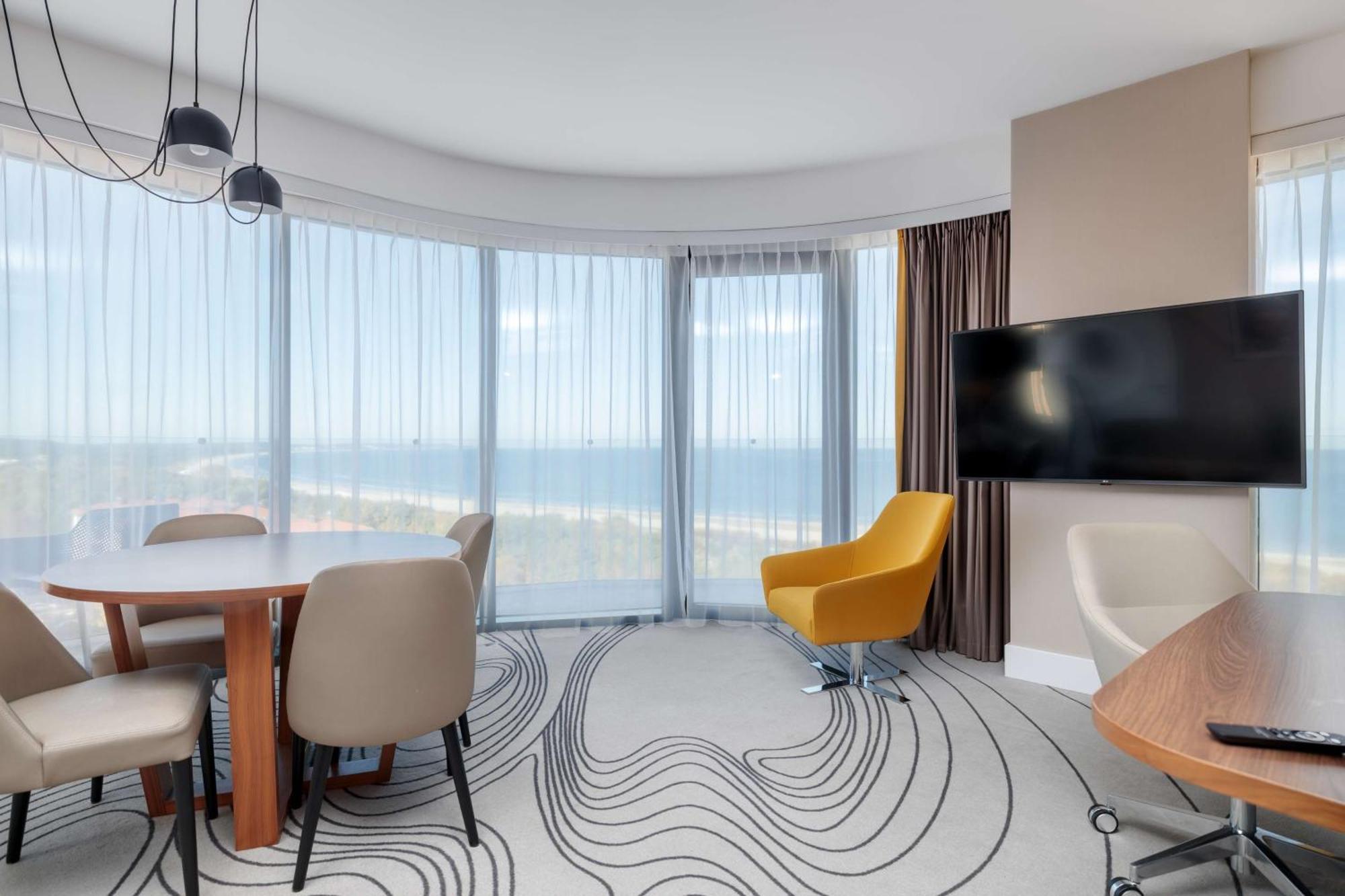 Radisson Blu Resort Swinoujscie Luaran gambar The photo shows a modern, stylish interior of a room with large windows that provide a panoramic view. The space features a round dining table surrounded by chairs, including a distinct yellow armchair. There is a flat-screen television mounted on th