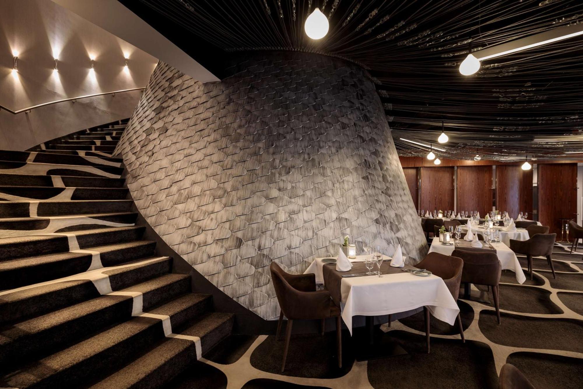 Radisson Blu Resort Swinoujscie Luaran gambar The photo shows the interior of a stylish restaurant. In the foreground, there are elegantly set tables with white tablecloths and dark chairs. A unique, curving stone wall adds a distinct architectural feature, and a grand staircase with a textured 