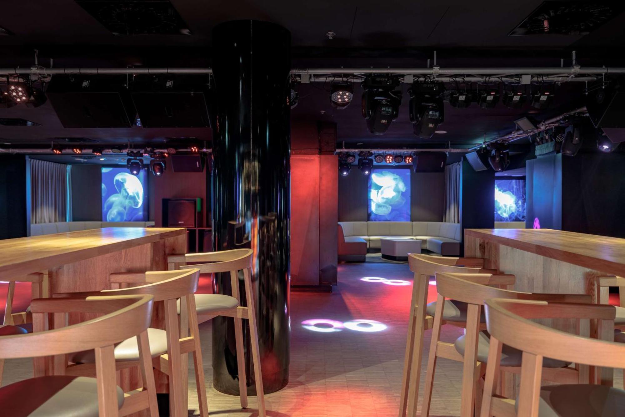 Radisson Blu Resort Swinoujscie Luaran gambar The photo shows an interior of a modern venue, likely a bar or club. The space features a central black column and light-colored wooden bar stools arranged around a bar area. On the walls, there are large screens displaying vibrant visuals, possibly 