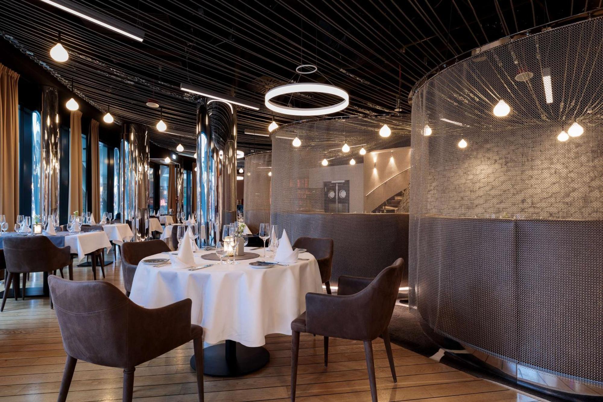 Radisson Blu Resort Swinoujscie Luaran gambar The image shows a modern restaurant interior that features a stylish design. There are several round tables with white tablecloths. The seating consists of brown, upholstered chairs. The space is illuminated by various hanging lights, including a dis